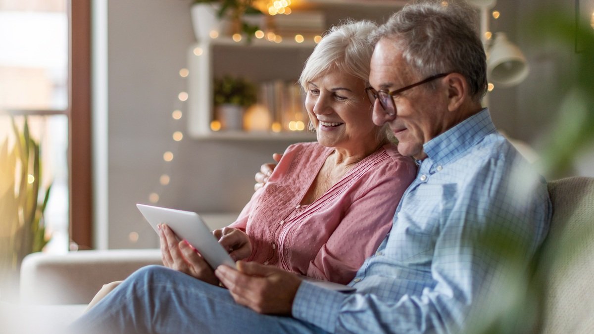 Immobilienverrentung - Seniorenpaar schaut gemeinsam auf Tablet im gemütlichen Wohnzimmer