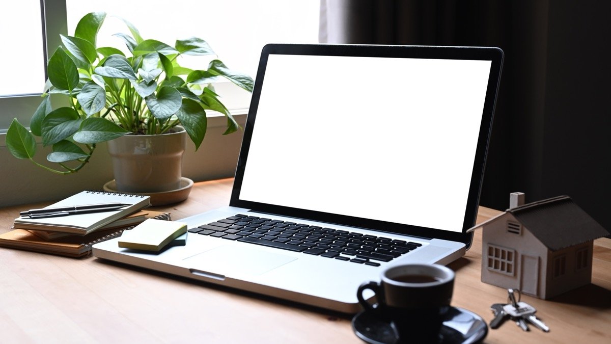 Mikroapartments kaufen - Laptop-Mockup am Fenster mit Hausmodell, Schlüsseln und Kaffeetasse