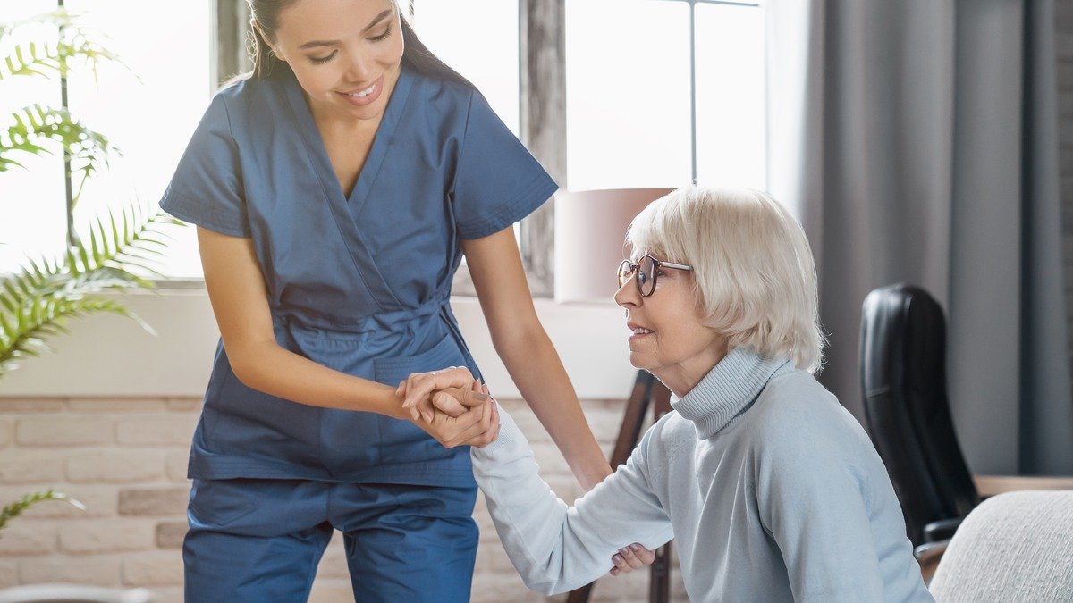 steigende_pflegekosten - Pflegekraft hilft Seniorin beim Aufstehen vom Sofa